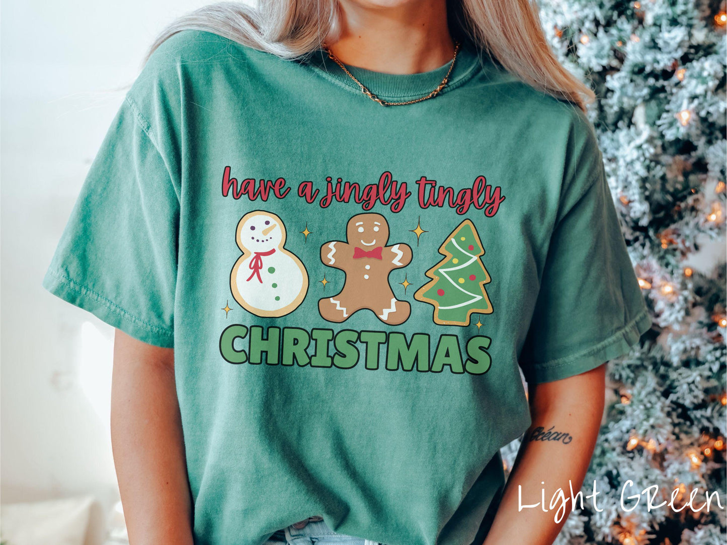 A vintage light green colored Christmas Comfort Colors t-shirt featuring a snowman iced cookie, a gingerbread man cookie, and a Christmas tree cookie. The text reads 'have a jingly tingly Christmas' in red and green font.