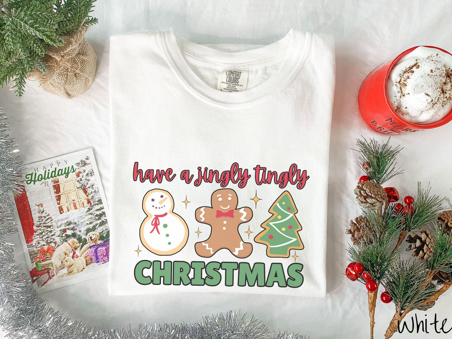 A vintage white colored Christmas Comfort Colors t-shirt featuring a snowman iced cookie, a gingerbread man cookie, and a Christmas tree cookie. The text reads 'have a jingly tingly Christmas' in red and green font.