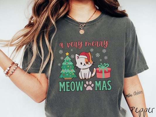 A vintage pepper colored Comfort Colors T-shirt with the text a very merry Christmas in red and green font, featuring a grey cat wearing a red Santa hat sitting between a Christmas tree and a present with white snowflakes hanging in the air.