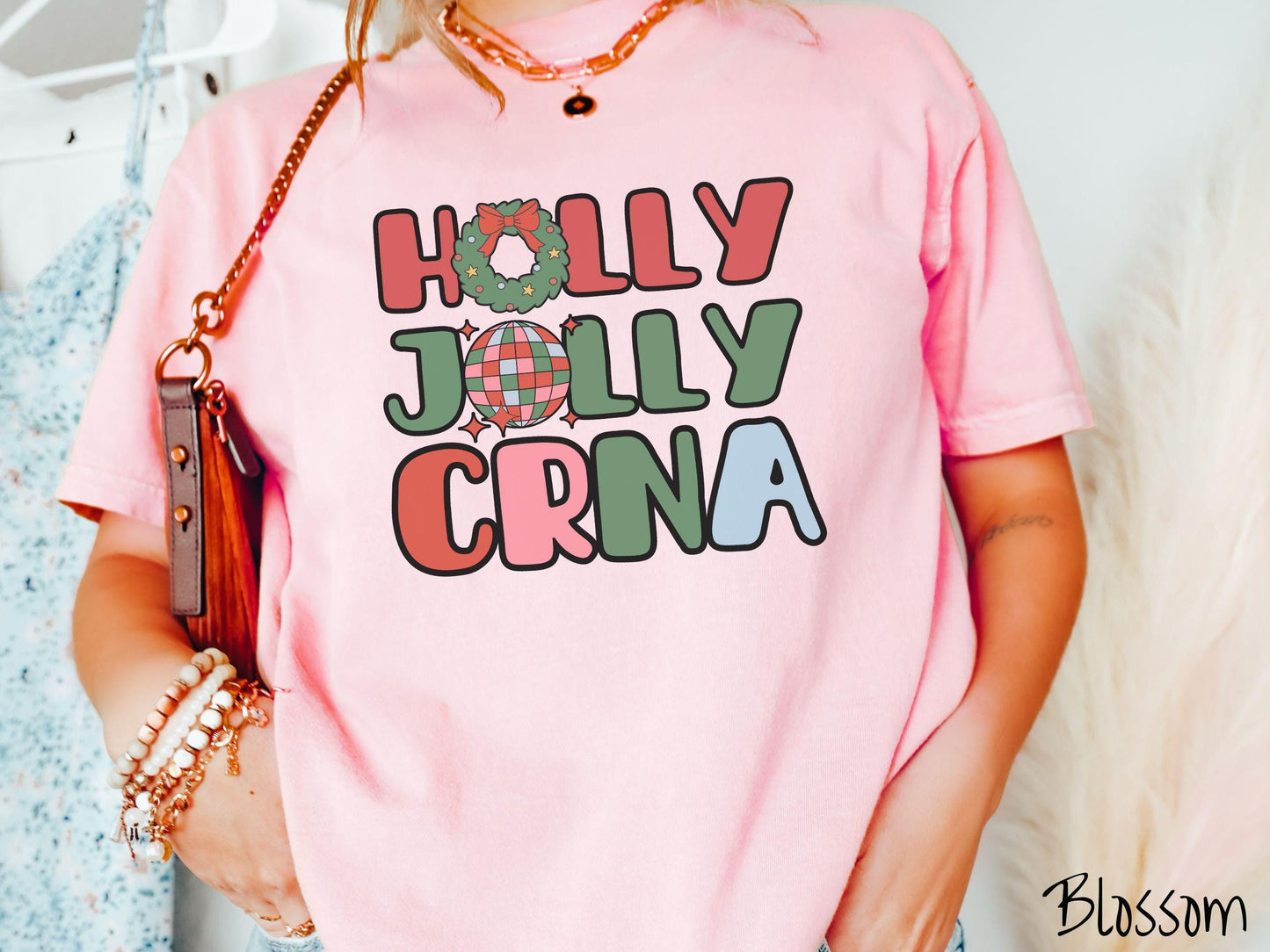 A woman wearing a cute, vintage blossom colored Comfort Colors T-shirt with the text Holly Jolly CRNA in red, green, pink, and blue font. The letter O in Holly is a Christmas wreath and the letter O in Jolly is a multi-colored disco ball.
