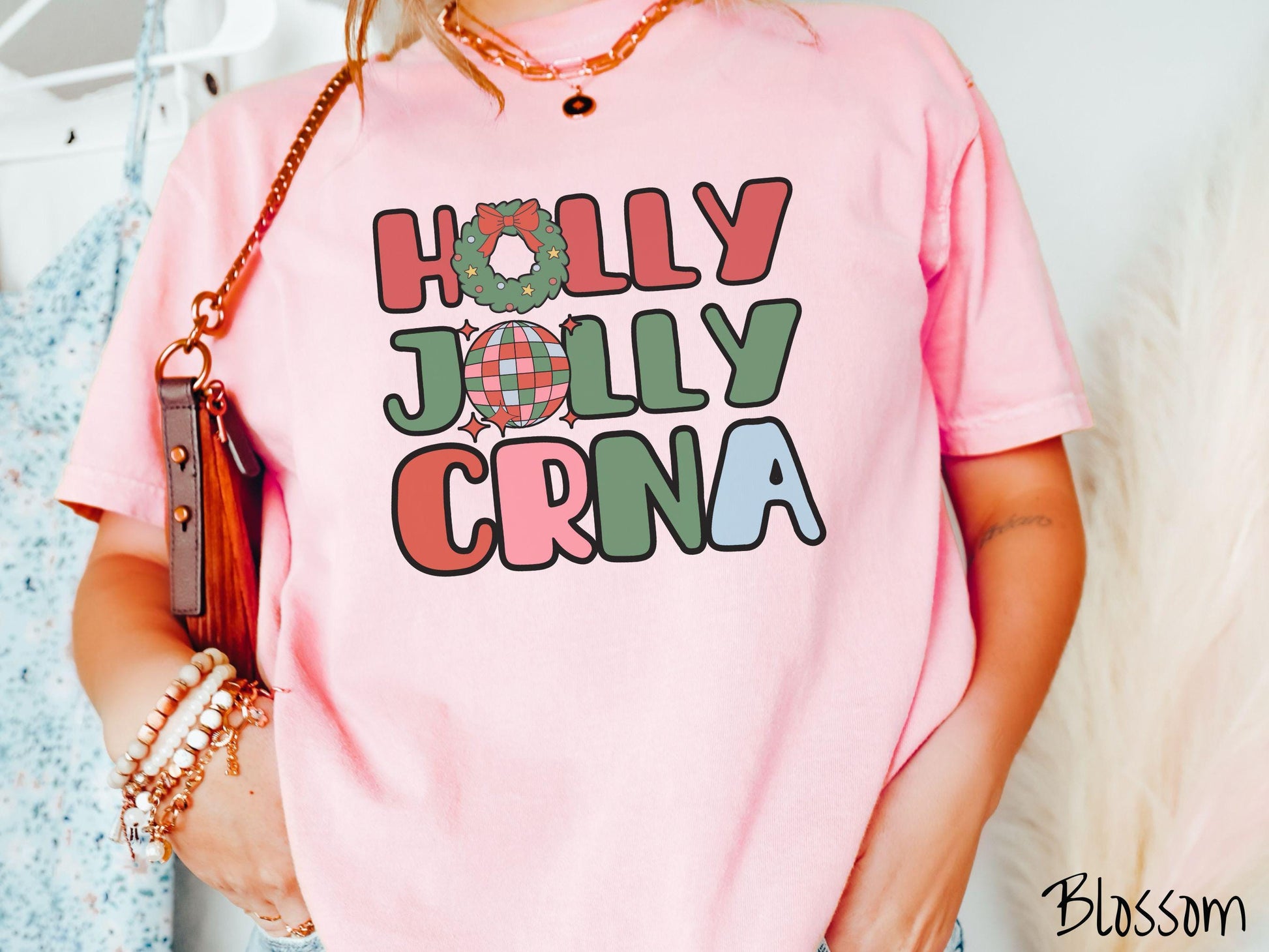 A woman wearing a cute, vintage blossom colored Comfort Colors T-shirt with the text Holly Jolly CRNA in red, green, pink, and blue font. The letter O in Holly is a Christmas wreath and the letter O in Jolly is a multi-colored disco ball.