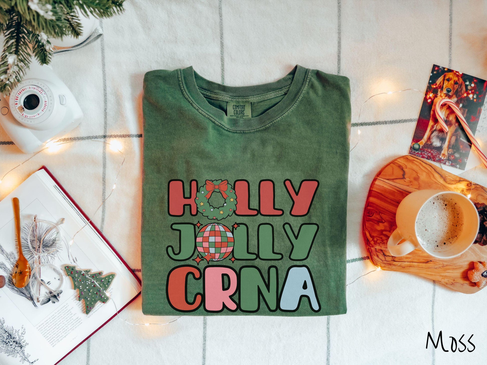 A cute, vintage moss colored Comfort Colors T-shirt with the text Holly Jolly CRNA in red, green, pink, and blue font. The letter O in Holly is a Christmas wreath and the letter O in Jolly is a multi-colored disco ball.