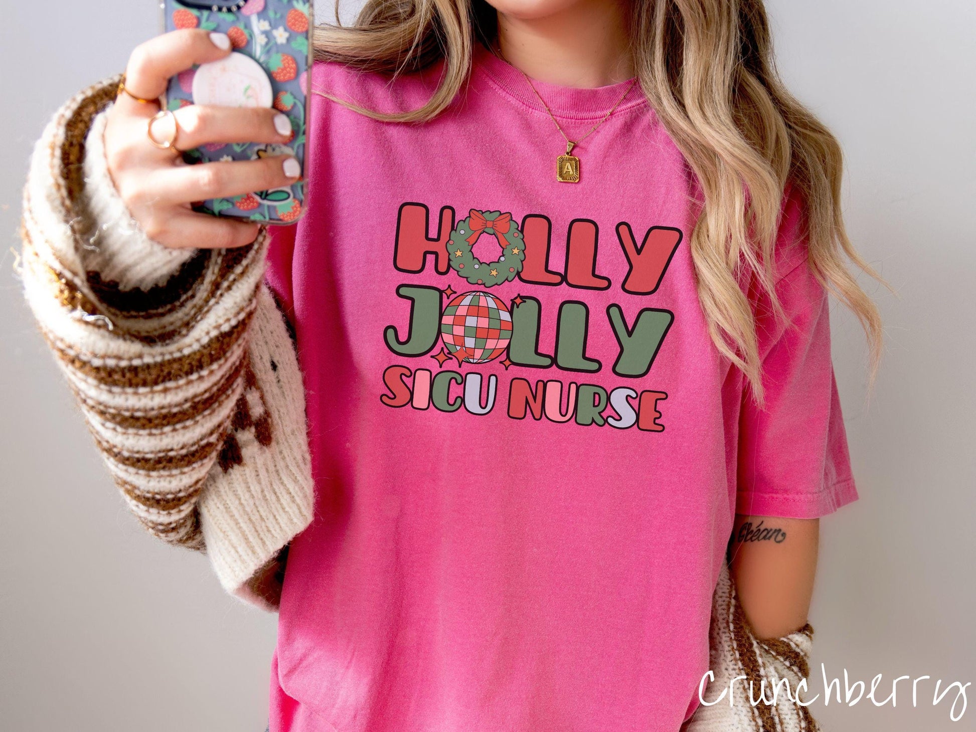 A woman wearing a cute, vintage crunchberry colored Comfort Colors T-shirt with the text Holly Jolly SICU Nurse in red, green, pink, and blue font. The letter O in Holly is a Christmas wreath and the letter O in Jolly is a multi-colored disco ball.