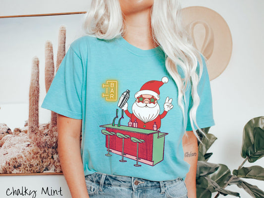 A vintage chalky mint colored Comfort Colors T-shirt features Santa Claus with red and green glasses mixing drinks and holding up a peace sign behind a red and green bar.