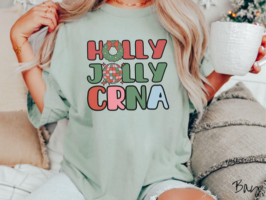 A woman wearing a cute, vintage bay colored Comfort Colors T-shirt with the text Holly Jolly CRNA in red, green, pink, and blue font. The letter O in Holly is a Christmas wreath and the letter O in Jolly is a multi-colored disco ball.