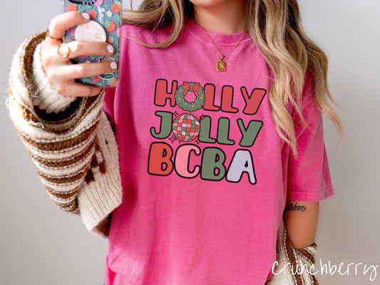 A woman wearing a cute, vintage crunchberry colored Comfort Colors T-shirt with the text Holly Jolly BCBA in red, green, pink, and blue font. The letter O in Holly is a Christmas wreath and the letter O in Jolly is a multi-colored disco ball.