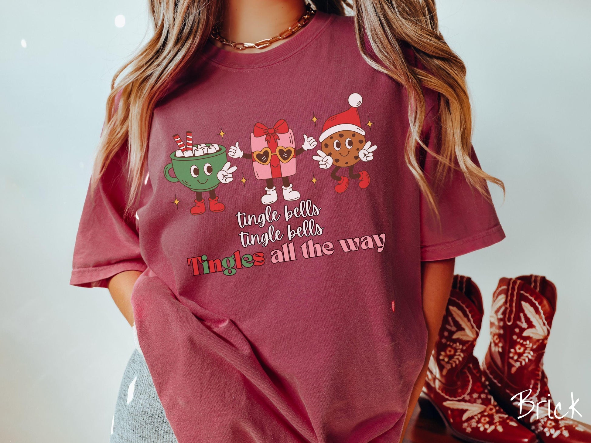 A cute, vintage brick colored Comfort Colors T-shirt with the text tnigle bells, tingle bells, tingles all the way. Above are a green cup of hot cocoa holding a peace sign, a red present holding two thumbs up, and a cookie holding up peace signs.