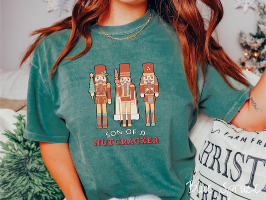 This vintage blue spruce colored Comfort Colors T-shirt features three red nutcracker figurines dressed in Christmas attire and one cracking a nut. Below is the text &#39;Son of a nutcracker&#39; in red and white font.