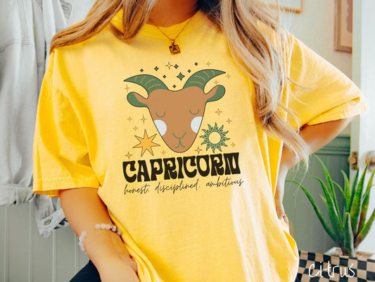 A vintage citrus colored Comfort Colors T-shirt features a large brown and green goat head surorunded by stars and the sun with the text Capricorn in large bold font and below that are the words honest, disciplined, ambitious in cursive font.