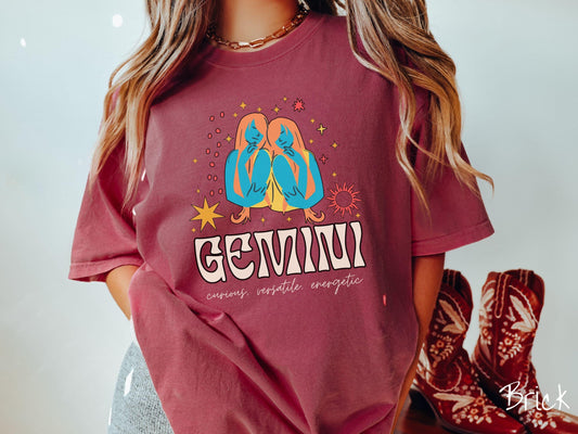 A vintage brick colored Comfort Colors T-shirt features the twins Castor and Pollux in blue and orange colors surrounded by stars and the sun with text Gemini in large bold font and below are the words curious, versatile, energetic in cursive font.