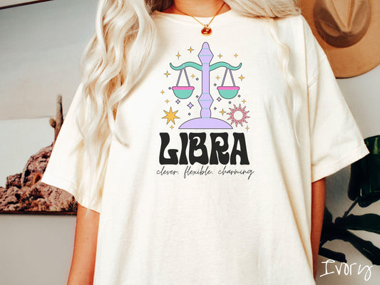 A vintage ivory colored Comfort Colors T-shirt features a large purple and green weight scale surrounded by stars and the sun with the text Libra in large bold font and below that are the words clever, flexible, charming in cursive font.