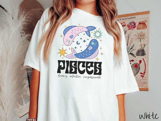 A vintage white colored Comfort Colors T-shirt features two large pink and blue fish surrounded by stars and the sun with the text Pisces in large bold font and below that are the words dreamy, intuitive, compassionate in cursive font.