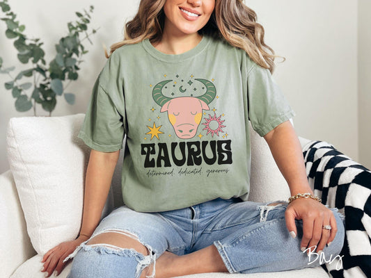 A vintage bay colored Comfort Colors T-shirt features a large pink and green bull surrounded by stars and the sun with the text Taurus in large bold font and below that are the words determined, dedicated, generous in cursive font.