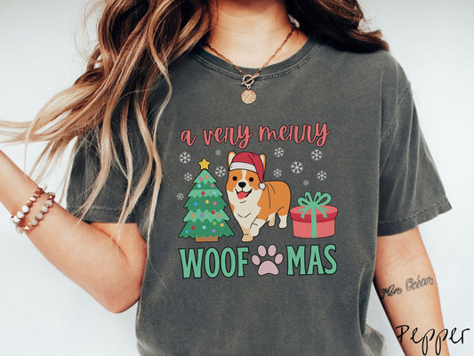 A vintage pepper colored Comfort Colors T-shirt with the text &#39;a very merry Woofmas&#39; in red and green font, featuring an orange corgi wearing a red Santa hat sitting between a Christmas tree and a present with snowflakes hanging in the air.