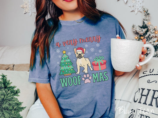 A vintage blue jean colored Comfort Colors T-shirt with the text &#39;a very merry Woofmas&#39; in red and green font, featuring a French Bulldog wearing a red Santa hat sitting between a Christmas tree and a present with white snowflakes hanging in the air.