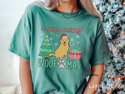 A vintage light green colored Comfort Colors T-shirt with the text &#39;a very merry Woofmas&#39; in red and green font, featuring a Golden Retriever wearing a red Santa hat sitting between a Christmas tree and a present with snowflakes hanging in the air.