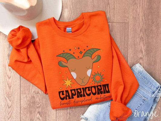 A vintage orange colored Gildan sweatshirt features a large brown and green goat head surrounded by stars and the sun with the text Capricorn in large bold font and below that are the words honest, disciplined, ambitious in cursive font.