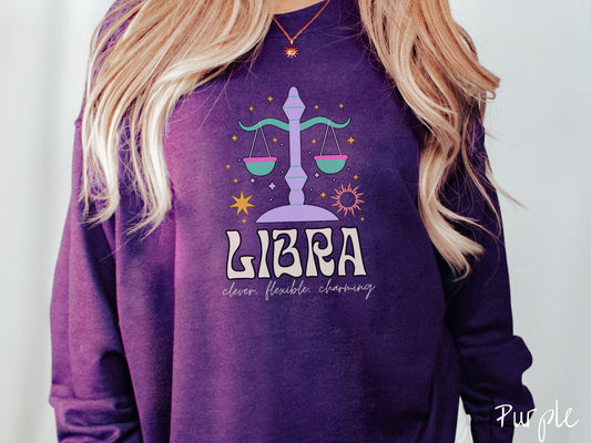 A vintage purple colored Gildan sweatshirt features a large purple and green weight scale surrounded by stars and the sun with the text Libra in large bold font and below that are the words clever, flexible, charming in cursive font.