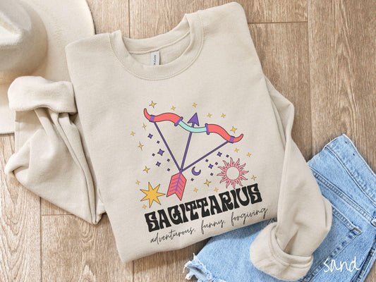 A vintage sand colored Gildan sweatshirt features a large red and purple bow and arrow surrounded by stars and the sun with the text Sagittarius in large bold font and below that are the words adventurous, funny, forgiving in cursive font.