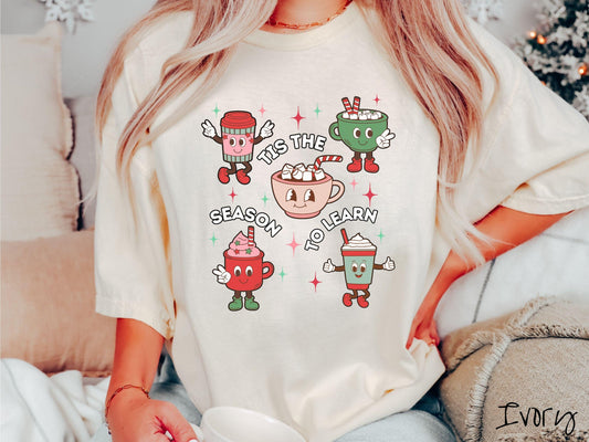 This vintage ivory colored Comfort Colors T-shirt features five coffee and hot cocoa cups with red and green festive decor surrounded by twinkling stars, and the text Tis the Season to Learn.