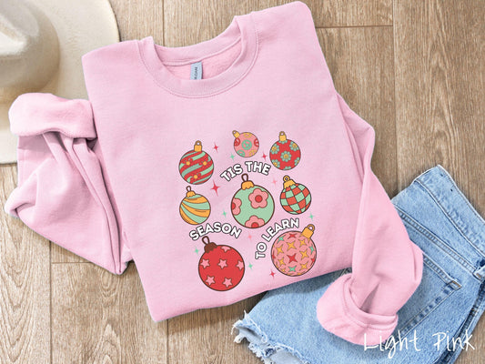 A vintage light pink colored Gildan sweatshirt features eight colorful Christmas tree ornaments arranged in a grid pattern and twinkling stars with the text Tis the Season to Learn.