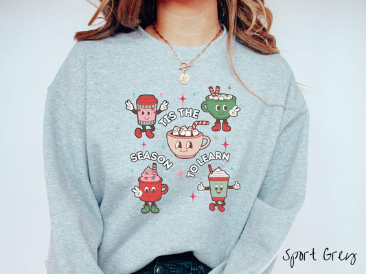 A vintage sport grey colored Gildan sweatshirt features five coffee and hot cocoa cups with red and green festive decor surrounded by twinkling stars, and the text Tis the Season to Learn.