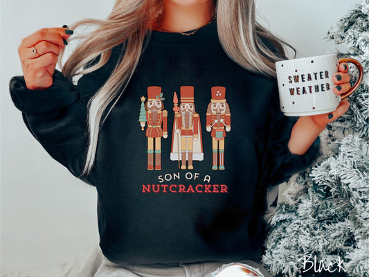 A vintage black colored Gildan sweatshirt features three red nutcracker figurines dressed in Christmas attire and one cracking a nut. Below is the text &#39;Son of a nutcracker&#39; in red and white font.