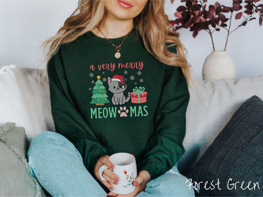 A vintage forest green colored Gildan sweatshirt with text &#39;a very merry Meowmas&#39; in red and green font, featuring a grey cat wearing a red Santa hat sitting between a decorated Christmas tree and a present with white snowflakes hanging in the air.