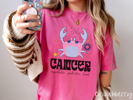 A vintage crunchberry colored Comfort Colors T-shirt features a large blue and pink crab surorunded by stars and the sun with the text Cancer in large bold font and below that ar the words empathetic, protective, and loyal in cursive font.