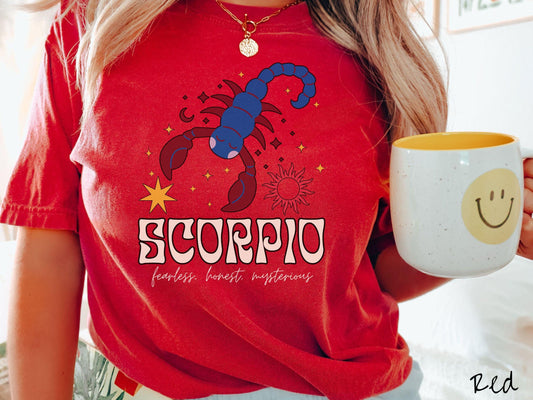 A vintage red colored Comfort Colors T-shirt features a large red and blue scorpion surrounded by stars and the sun with the text Scorpio in large bold font and below that are the words fearless, honest, mysterious in cursive font.