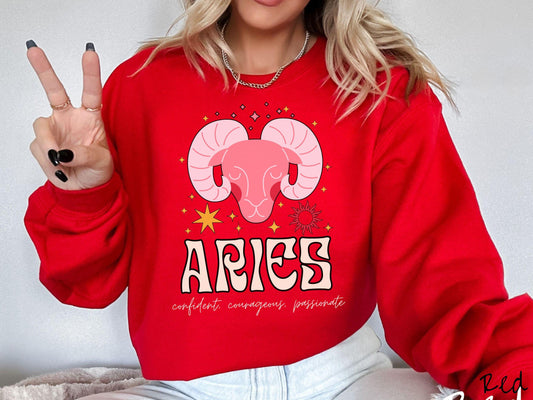 A vintage red colored Comfort Colors sweatshirt features a large red ram head surrounded by stars and the sun, with the text Aries in large bold font and below that are the words confident, courageous, passionate in cursive font.