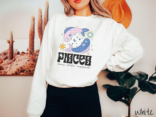 A vintage white colored Gildan sweatshirt features two large pink and blue fish surrounded by stars and the sun with the text Pisces in large bold font and below that are the words dreamy, intuitive, compassionate in cursive font.