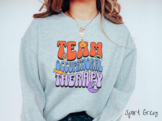 A vintage, sport grey colored Gildan sweatshirt with the text &quot;Team Occupational Therapy&quot; surrounded by playful elements like a chocolate ice cream cone, a blazing flame, a colorful rainbow, a piece of candy, and a serene moon.