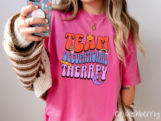 A vintage, crunchberry colored Comfort Colors t-shirt with the text &quot;Team Occupational Therapy&quot; surrounded by playful elements like a chocolate ice cream cone, a blazing flame, a colorful rainbow, a piece of candy, and a serene moon.