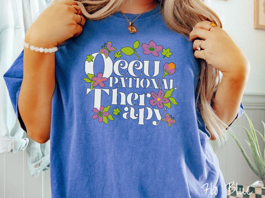 A vintage, flo blue colored Comfort Colors t-shirt with the text Occupational Therapy and featuring green leaves, purple and orange flowers, and colorful butterflies in the background.