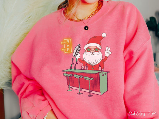 A vintage safety pink colored Gildan sweatshirt features Santa Claus with red and green glasses mixing drinks and holding up a peace sign behind a red and green bar.