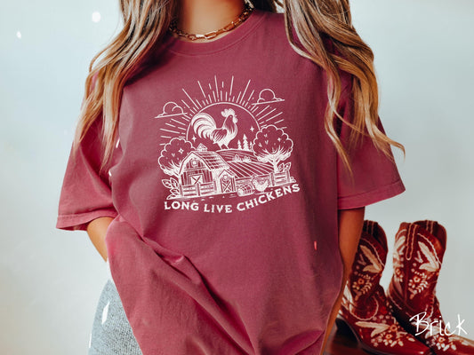 A vintage brick colored Comfort Colors t-shirt featuring a design of a farm with a fence, trees, and plants scattered along the property, a huge chicken in the background at the top of a pasture in front of the sun, and text Long Live Chickens.