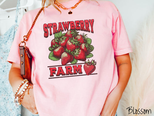 A vintage blossom colored Comfort Colors t-shirt featuring the text Strawberry Farm in red font with a bundle of red strawberries in between the text.