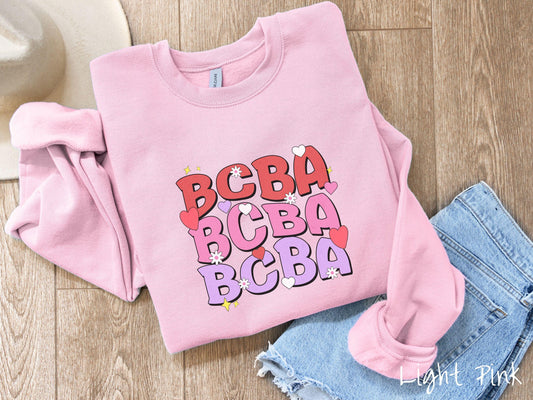A vibrant Gildan Sweatshirt with the text BCBA written three times vertically, accompanied by tiny hearts, stars, and flowers. Perfect for Board Certified Behavior Analysts, this tee is a great Valentine&#39;s Day gift to show appreciation and love.