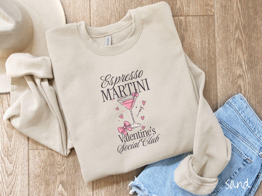 A stylish Gildan sweatshirt featuring the phrase &quot;Espresso Martini&quot; above a martini glass filled with pink martini and a pink bow-topped toothpick. The words &quot;Valentine&#39;s Social Club&quot; appear below with pink bows and hearts throughout the design.