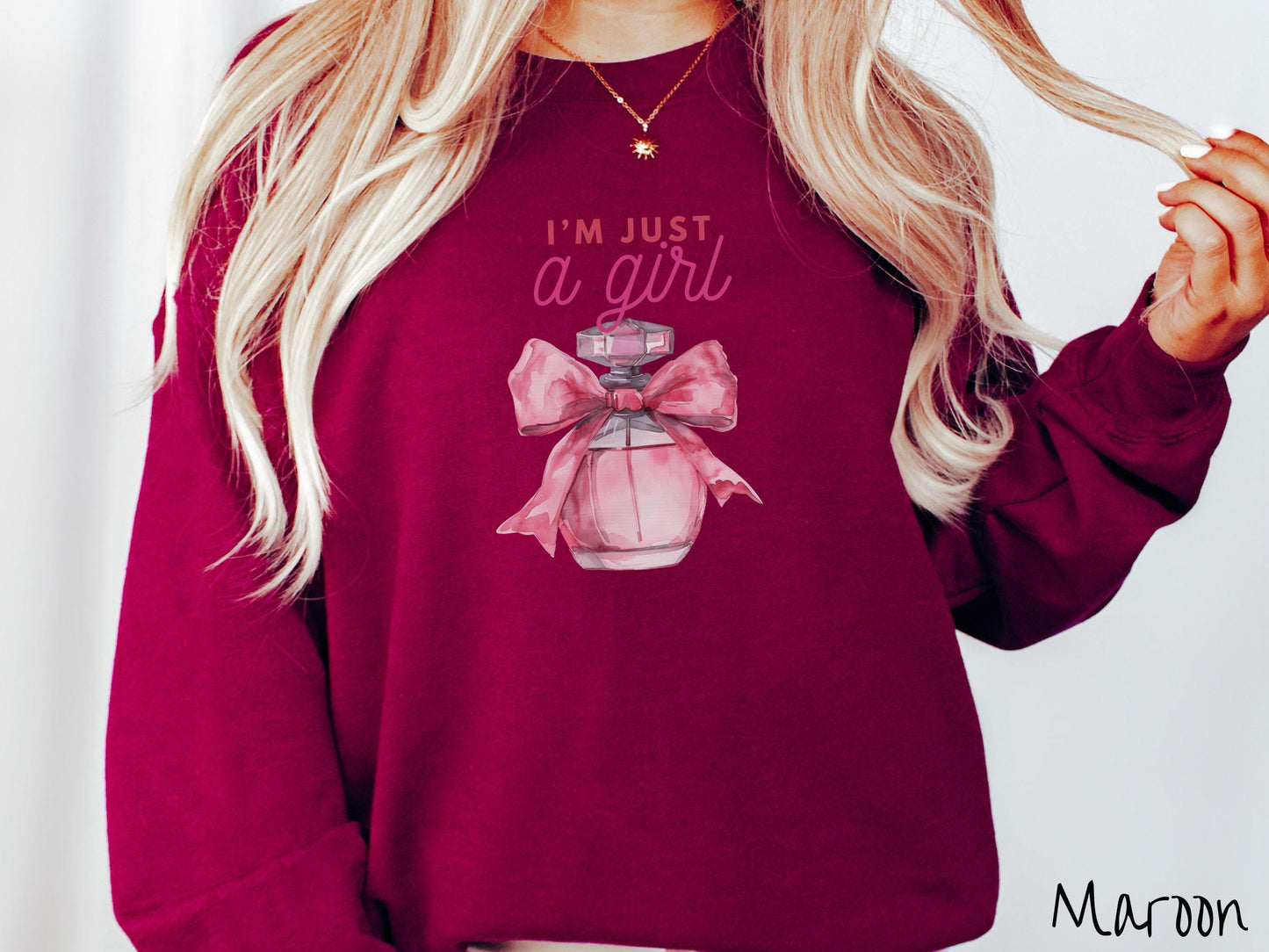 A cozy Gildan sweatshirt featuring the phrase &quot;I&#39;m Just a Girl&quot; in pink font above a bottle of pink perfume tied with a pink bow. Perfect for those colder months, this sweatshirt provides warmth and comfort with its classic fit and crew neckline.