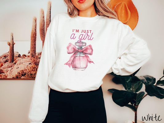 A cozy Gildan sweatshirt featuring the phrase &quot;I&#39;m Just a Girl&quot; in pink font above a bottle of pink perfume tied with a pink bow. Perfect for those colder months, this sweatshirt provides warmth and comfort with its classic fit and crew neckline.
