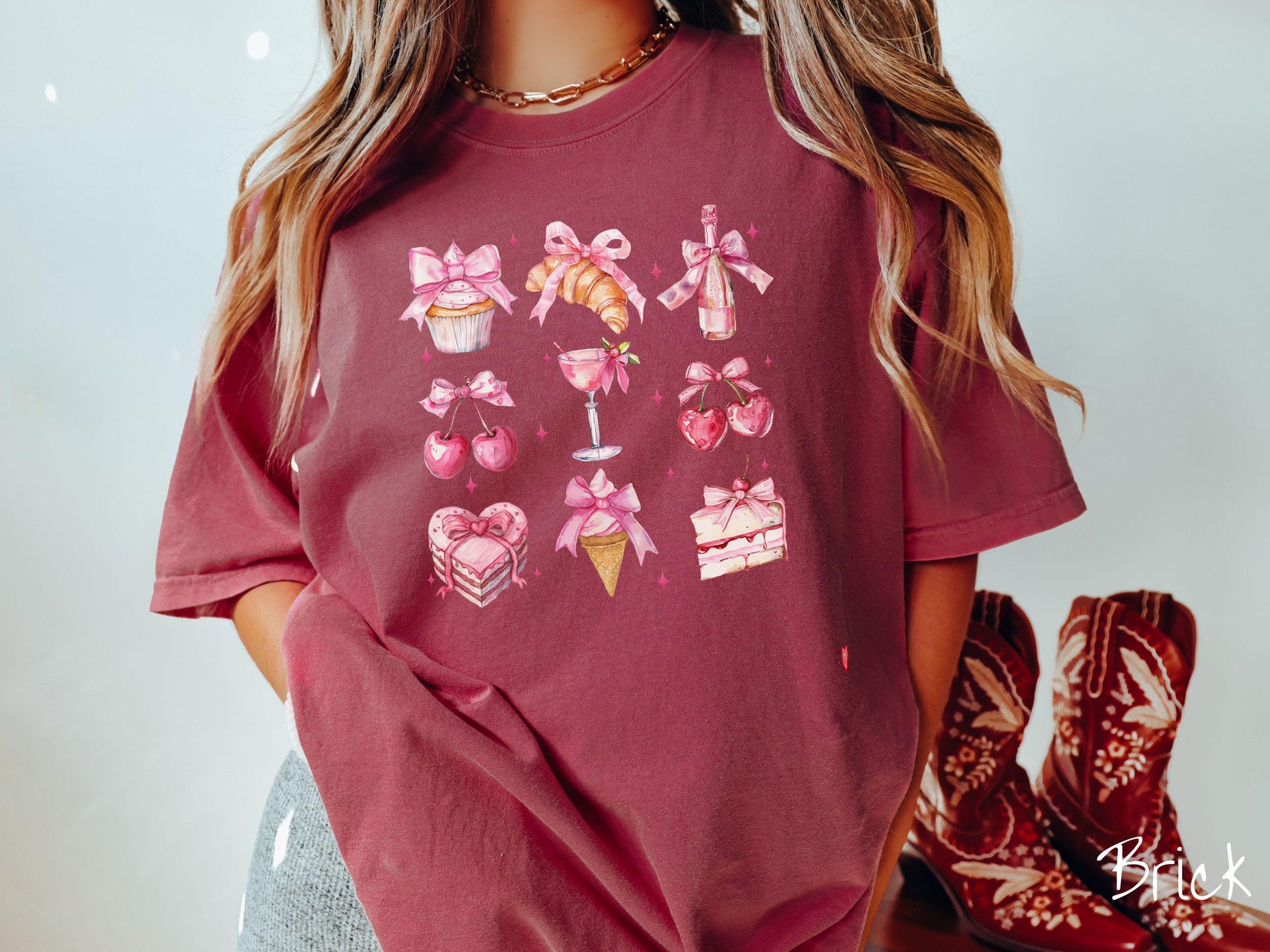 A stylish coquette Comfort Colors T-shirt featuring a grid of assorted treats with pink ribbons like a cupcake, a croissant, a champaigne bottle, cherries, a pink martini, a pink cake, and a pink ince cream cone, along with twinkling pink stars.