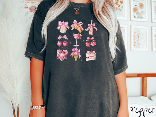 A stylish coquette Comfort Colors T-shirt featuring a grid of assorted treats with pink ribbons like a cupcake, a croissant, a champaigne bottle, cherries, a pink martini, a pink cake, and a pink ince cream cone, along with twinkling pink stars.