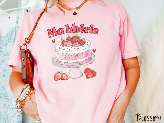 A Comfort Colors T-shirt featuring the phrase &quot;Ma Bherie&quot; is printed in bold red font above a charming illustration of a red-and-white strawberry cake displayed on a plate, topped with fresh strawberries and surrounded by even more berries.