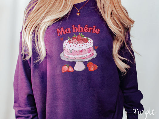 A Gildan sweatshirt featuring the phrase &quot;Ma Bherie&quot; is printed in bold red font above a charming illustration of a red-and-white strawberry cake displayed on a plate, topped with fresh strawberries and surrounded by even more berries.