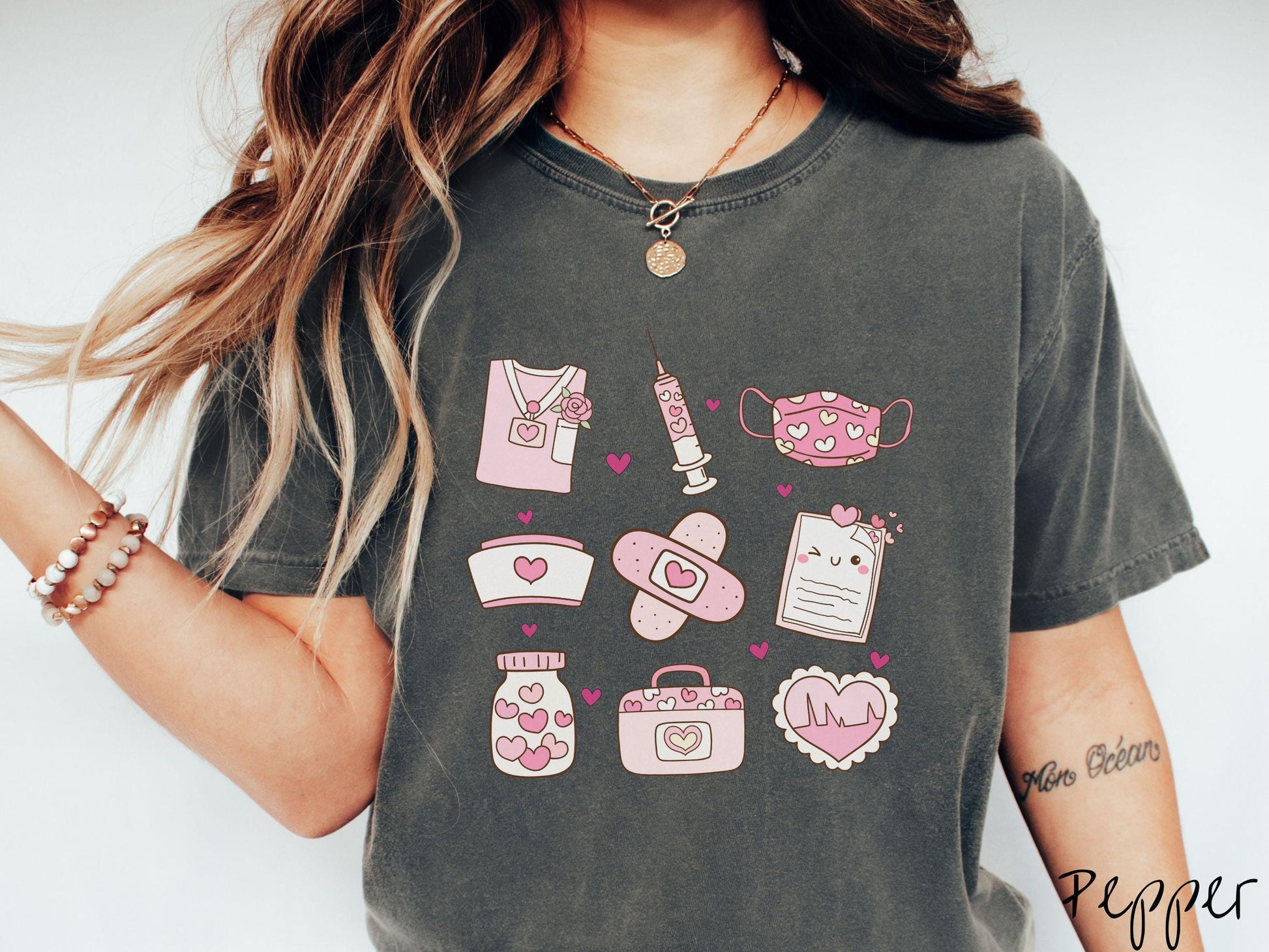 A Comfort Colors T-shirt featuring a 3x3 grid of nursing-themed illustrations adorned with heart designs like nursing scrubs, syringes, a face mask, a nurse cap, bandages, medical records, vitamins, a medical bag, and a heart-shaped EKG graph.