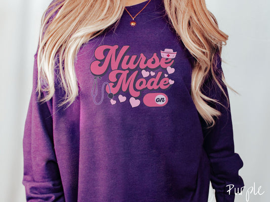 A stylish Gildan sweatshirt design features the text &quot;Nurse Mode&quot; in bold pink font with a pink nurse&#39;s cap placed over the letter E, and a stethoscope positioned beneath the text, alongside an &quot;on&quot; button widget set to the on position.