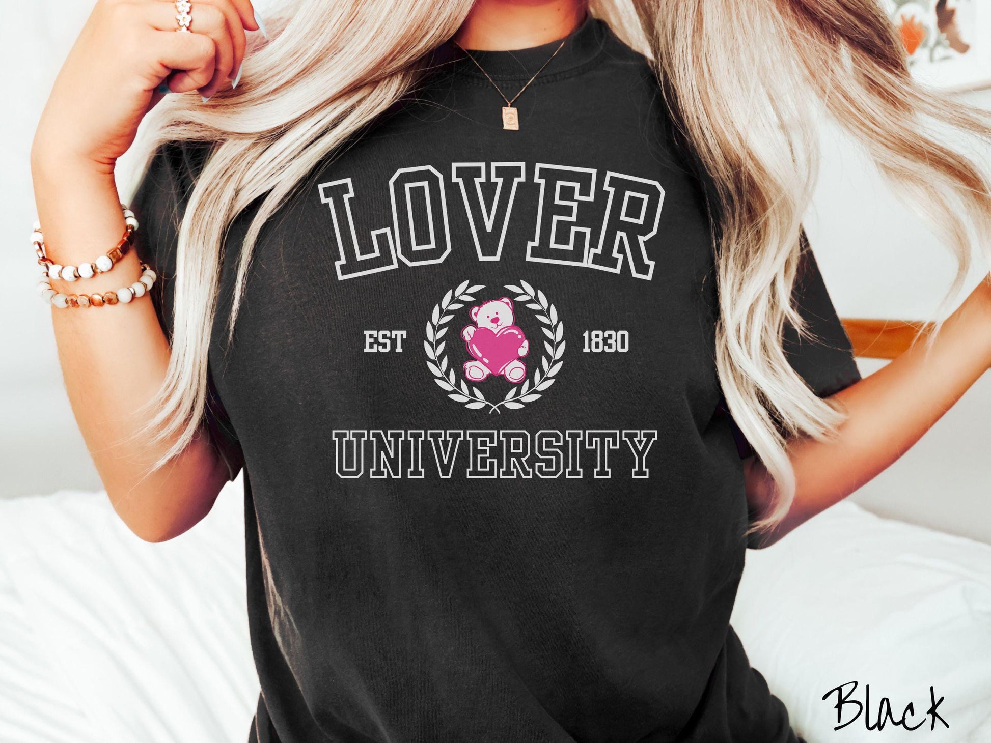 A vintage Comfort Colors t-shirt featuring a university logo-inspired design showcasing the text “Lover University” in collegiate-style font. Two pink laurel leaves frame a pink teddy bear hugging a heart with the text “EST. 1830” in pink font.