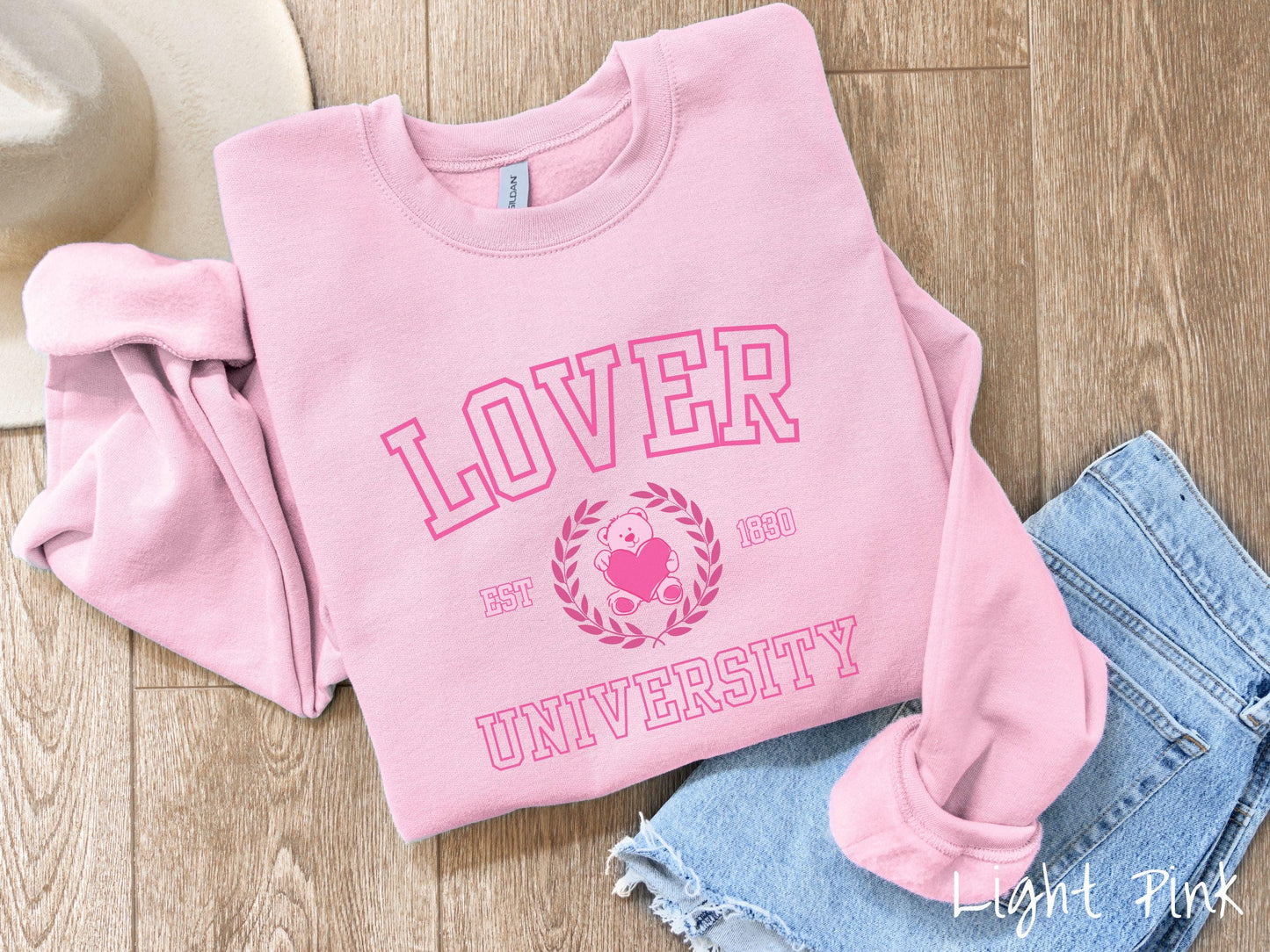A vintage Gildan sweatshirt featuring a university logo-inspired design showcasing the text “Lover University” in collegiate-style font. Two pink laurel leaves frame a pink teddy bear hugging a heart with the text “EST. 1830” in pink font.